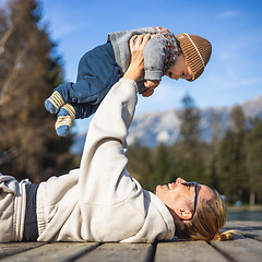 Image showing Happy family. Young mother playing with her baby boy infant oudoors on sunny autumn day. Portrait of mom and little son on wooden platform by lake. Positive human emotions, feelings, joy.