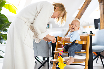 Image showing Happy infant sitting and playing with his toy in traditional scandinavian designer wooden high chair in modern bright atic home suppervised by his mother.