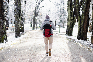 Image showing Rear view of sporty father carrying his infant son wearing winter jumpsuit and cap in backpack carrier walking in city park in winter