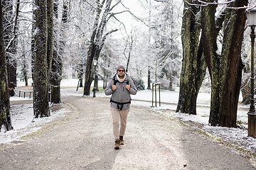 Image showing Sporty father carrying his infant son wearing winter jumpsuit and cap in backpack carrier walking in city park in winter