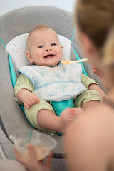 Image showing Mother spoon feeding her baby boy infant child in baby chair with fruit puree. Baby solid food introduction concept.