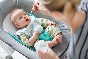 Image showing Mother spoon feeding her baby boy infant child in baby chair with fruit puree. Baby solid food introduction concept.