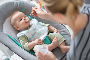 Image showing Mother spoon feeding her baby boy infant child in baby chair with fruit puree. Baby solid food introduction concept.