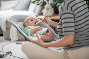 Image showing Mother spoon feeding her baby boy infant child in baby chair with fruit puree. Baby solid food introduction concept.