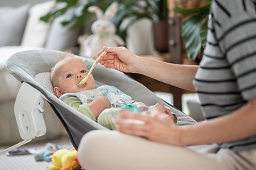 Image showing Mother spoon feeding her baby boy infant child in baby chair with fruit puree. Baby solid food introduction concept.