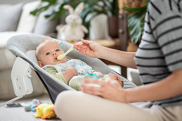 Image showing Mother spoon feeding her baby boy infant child in baby chair with fruit puree. Baby solid food introduction concept.