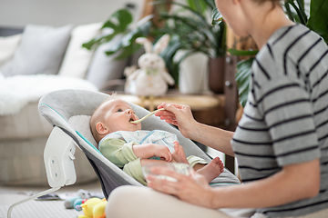 Image showing Mother spoon feeding her baby boy infant child in baby chair with fruit puree. Baby solid food introduction concept.