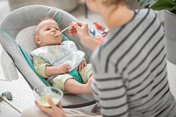 Image showing Mother spoon feeding her baby boy infant child in baby chair with fruit puree. Baby solid food introduction concept.