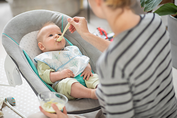 Image showing Mother spoon feeding her baby boy infant child in baby chair with fruit puree. Baby solid food introduction concept.