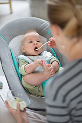 Image showing Mother spoon feeding her baby boy infant child in baby chair with fruit puree. Baby solid food introduction concept.