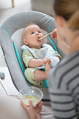 Image showing Mother spoon feeding her baby boy infant child in baby chair with fruit puree. Baby solid food introduction concept.