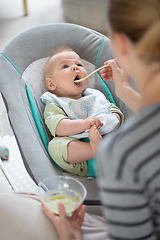 Image showing Mother spoon feeding her baby boy infant child in baby chair with fruit puree. Baby solid food introduction concept.