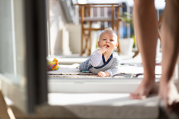 Image showing Cute little infant baby boy playing with toys outdoors at the patio in summer being supervised by her mother seen in the background.