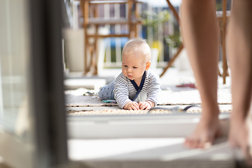 Image showing Cute little infant baby boy playing with toys outdoors at the patio in summer being supervised by her mother seen in the background.