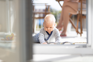 Image showing Cute little infant baby boy playing with toys outdoors at the patio in summer being supervised by her mother seen in the background.