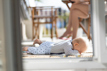 Image showing Cute little infant baby boy sulking while playing with toys outdoors at the patio in summer being supervised by her mother seen in the background.