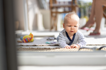 Image showing Cute little infant baby boy playing with toys outdoors at the patio in summer being supervised by her mother seen in the background.