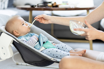 Image showing Mother spoon feeding her baby boy infant child in baby chair with fruit puree. Baby solid food introduction concept.