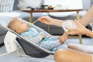 Image showing Mother spoon feeding her baby boy infant child in baby chair with fruit puree. Baby solid food introduction concept.