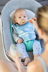 Image showing Mother spoon feeding her baby boy infant child in baby chair with fruit puree. Baby solid food introduction concept.