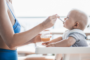 Image showing Mother spoon feeding her baby boy child in baby chair with fruit puree on a porch on summer vacations. Baby solid food introduction concept.