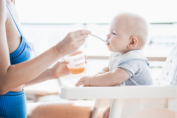 Image showing Mother spoon feeding her baby boy child in baby chair with fruit puree on a porch on summer vacations. Baby solid food introduction concept.