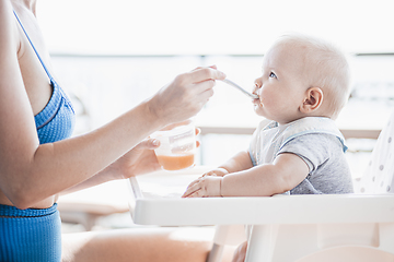 Image showing Mother spoon feeding her baby boy child in baby chair with fruit puree on a porch on summer vacations. Baby solid food introduction concept.