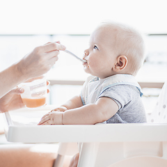 Image showing Mother spoon feeding her baby boy child in baby chair with fruit puree on a porch on summer vacations. Baby solid food introduction concept.