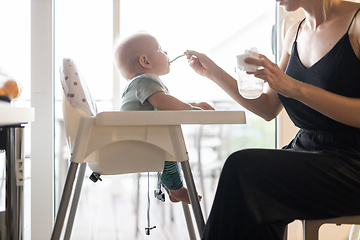 Image showing Mother spoon feeding her baby boy child in baby chair with fruit puree in kitchen at home. Baby solid food introduction concept.