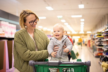 Image showing Mother pushing shopping cart with her infant baby boy child down department aisle in supermarket grocery store. Shopping with kids concept.