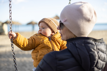 Image showing Mother pushing her infant baby boy child on a swing on sandy beach playground outdoors on nice sunny cold winter day in Malaga, Spain.
