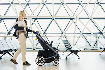 Image showing Mother carying his infant baby boy child, pushing stroller at airport departure terminal moving to boarding gates to board an airplane. Family travel with baby concept.