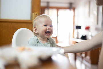 Image showing Adorable cheerful happy infant baby boy child smiling while sitting in high chair at the dining table in kitchen at home beeing spoon fed by his mother