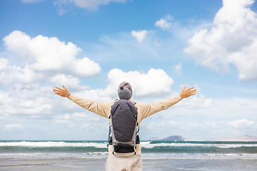 Image showing Young father rising hands to the sky while enjoying pure nature carrying his infant baby boy son in backpack on windy sandy beach of Famara, Lanzarote island, Spain. Family travel concept.