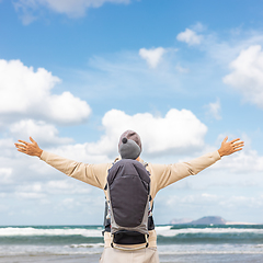 Image showing Young father rising hands to the sky while enjoying pure nature carrying his infant baby boy son in backpack on windy sandy beach of Famara, Lanzarote island, Spain. Family travel concept.