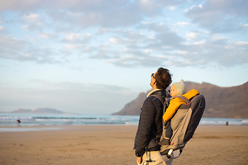 Image showing Young father rising hands to the sky while enjoying pure nature carrying his infant baby boy son in backpack on windy sandy beach of Famara, Lanzarote island, Spain at sunset. Family travel concept.