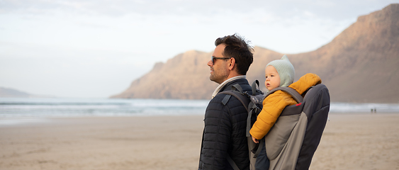 Image showing Father enjoying pure nature carrying his infant baby boy son in backpack on windy sandy beach of Famara, Lanzarote island, Spain at sunset. Family travel concept.