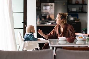 Image showing Cheerful mother wearing bathrope spoon feeding her infant baby boy child sitting in high chair at the dining table in kitchen at home in the morning.