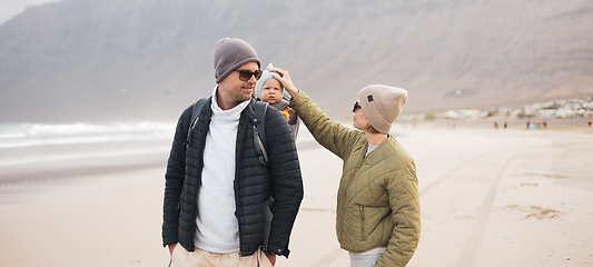 Image showing Young father carrying his infant baby boy son in backpack on windy sandy beach. Family travel and winter vacation concept.