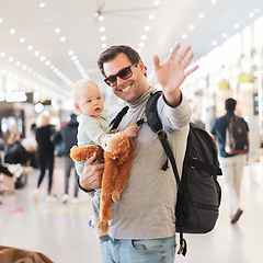 Image showing Father traveling with child, holding his infant baby boy at airport terminal waiting to board a plane waving goodby. Travel with kids concept.