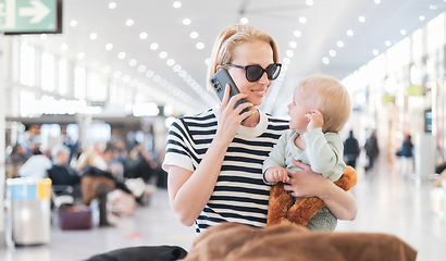 Image showing Mother talking on mobile phone while traveling with child, holding his infant baby boy at airport terminal waiting to board a plane. Travel with kids concept.