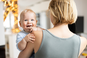 Image showing Blonde mother holding her adorable cheerful little infant baby boy. Cute infant child looking in camera and smiling at home.