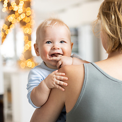 Image showing Blonde mother holding her adorable cheerful little infant baby boy. Cute infant child looking in camera and smiling at home.