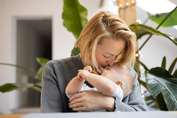 Image showing Portrait of young mother cuddling and kissing her adorable little child while sitting at the table at home. Sensory stimulation for baby development