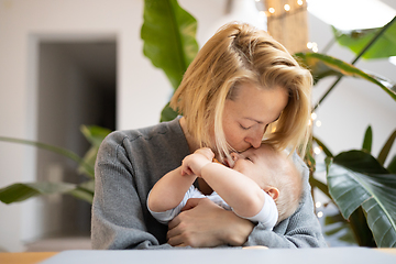 Image showing Portrait of young mother cuddling and kissing her adorable little child while sitting at the table at home. Sensory stimulation for baby development