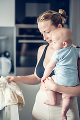 Image showing Woman wiping kitchen sink with a cloth after finishing washing the dishes while holding four months old baby boy in her hands