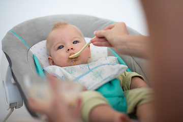 Image showing Mother spoon feeding her baby boy infant child in baby chair with fruit puree. Baby solid food introduction concept.