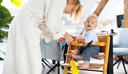 Image showing Happy infant sitting and playing with his toy in traditional scandinavian designer wooden high chair in modern bright atic home suppervised by his mother.