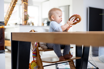 Image showing Happy infant sitting at dining table and playing with his toy in traditional scandinavian designer wooden high chair in modern bright atic home. Cute baby playing with toys