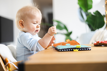 Image showing Happy infant sitting at dining table and playing with his toy in traditional scandinavian designer wooden high chair in modern bright atic home. Cute baby playing with toys